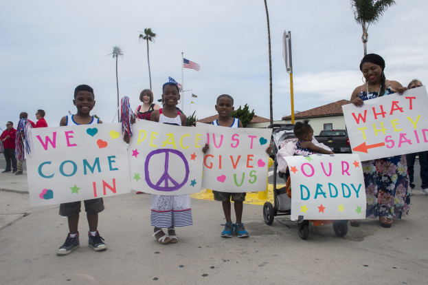 Eine Gruppe von Kindern hält Schilder auf einer Straße vor einem Gebäude, während eine Frau einen Kinderwagen schiebt, in dem ein Baby liegt.