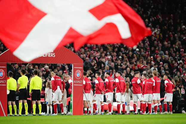 Gruppe von Menschen auf einem Fußballfeld mit einer roten und weißen Flagge im Vordergrund, einem Bogen mit der Aufschrift "Bayern München vs Bayern Munich Betting Tips & Preview" im Hintergrund und einer großen Stadionmenge.