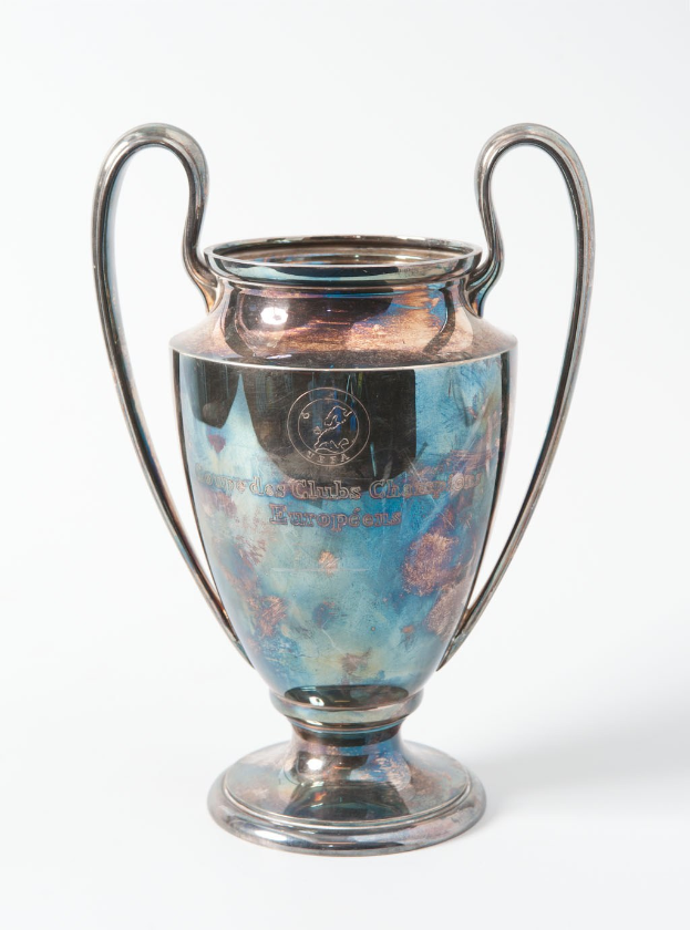 Der UEFA-Champions-League-Pokal, ein stählerner Becher mit zwei Griffen und einem blauen und silbernen Farbschema, der mit 'UEFA Champions League' beschriftet ist und gegen einen weißen Hintergrund gehalten wird.