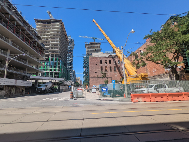 Baustelle mit einem Kran, umgeben von Gebäuden, Bäumen, Straßeninfrastruktur, Fahrzeugen und einem klaren blauen Himmel.