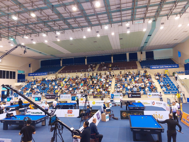 Ein großer Raum mit mehreren Poolbillardtischen umgeben von sitzenden Spielern, Fenster an der linken Wand, Deckenbeleuchtung und eine Kameraaufstellung, die auf ein professionelles Poolbillardturnier hinweist.