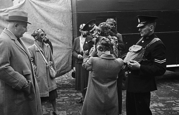 Schwarzes und weißes Bild einer Gruppe von Menschen in Mänteln und Mützen mit Gasmasken, die vor einem Zelt mit einem Fahrzeug im Hintergrund stehen.