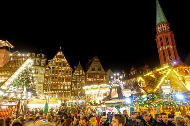 Ein lebhafter Weihnachtsmarkt in Nürnberg, Deutschland, mit Menschen um geschmückte Stände, Gebäuden mit Fenstern und einem Kirchturm unter einem dunklen Himmel.