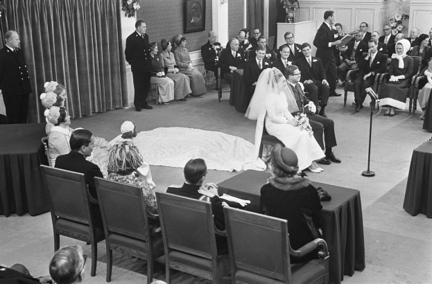 Schwarzes und weißes Foto einer Hochzeitszeremonie mit sitzenden und stehenden Menschen, einem Mann, der in ein Mikrofon spricht, und einer Frau, die einen Blumenstrauß hält.