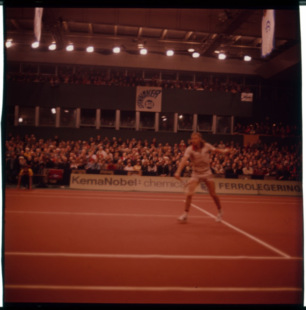 Ein Mann spielt Tennis auf einem Sandplatz unter hellen Scheinwerfern, umgeben von Zuschauern auf Stühlen und Bannern.