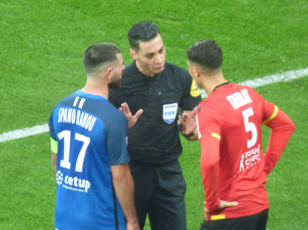 Schiedsrichter in schwarzem Hemd und Hose, der mit zwei Männern in blauer und roter Kleidung auf einem Fußballfeld spricht, alle tragen Brillen.