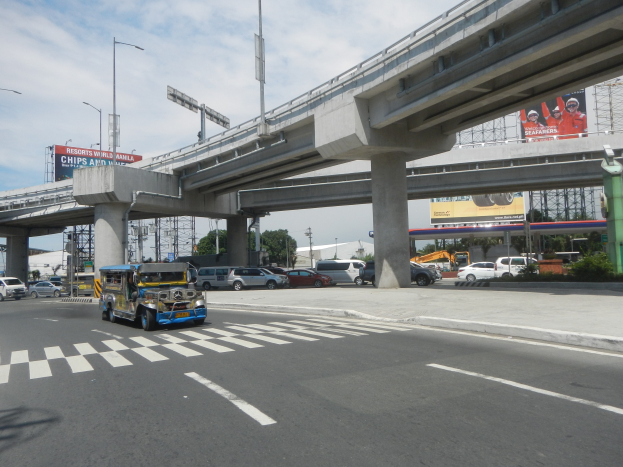Vielbefahrene Hauptstraße mit Fahrzeugen, Zebrastreifen, Überführung mit Pfeilern, Laternen, Schilder, Bäume, Gebäude und bewölkter Himmel im Hintergrund.
