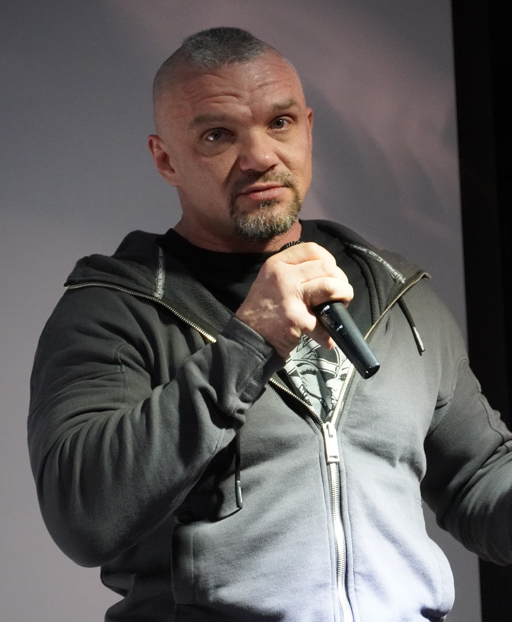 Ein Mann in einer schwarzen Kapuzenjacke und einem schwarzen Anzug mit einem weißen Hemd, der ein Mikrofon hält und selbstbewusst mit glattem Haar vor einer Wand steht.
