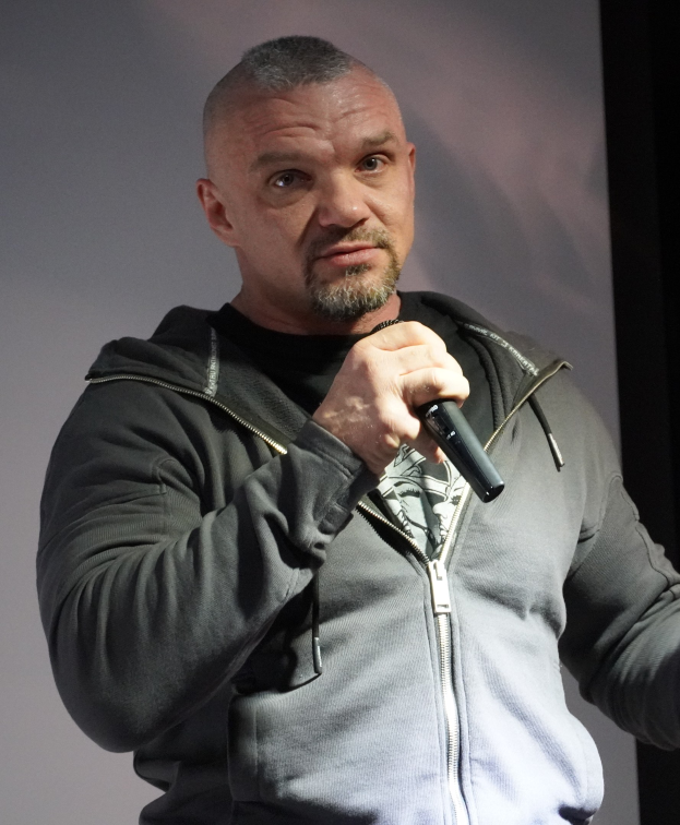 Ein Mann in einer schwarzen Kapuzenjacke und einem schwarzen Anzug mit einem weißen Hemd, der ein Mikrofon hält und selbstbewusst mit glattem Haar vor einer Wand steht.