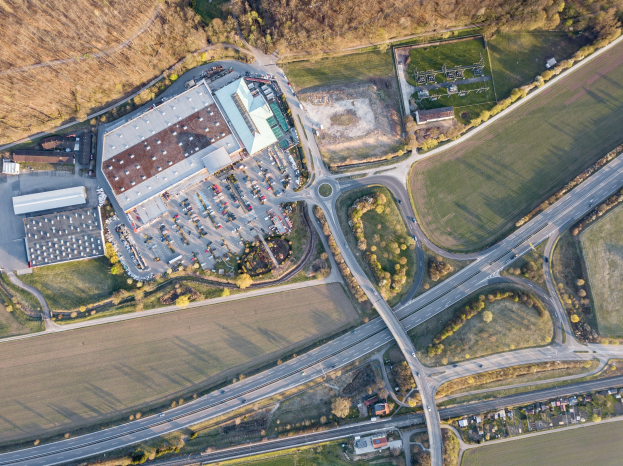 Luftaufnahme eines großen Gebäudes mit Firmenlogo, umgeben von Straßen, Fahrzeugen, Bäumen und Gras.