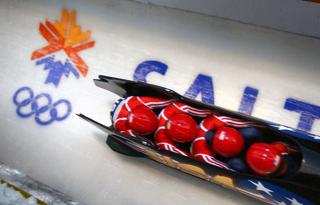 Ein Rodler in einem Schlitten auf einer olympischen Bahn, mit einer dekorierten Wand und Text im Hintergrund.