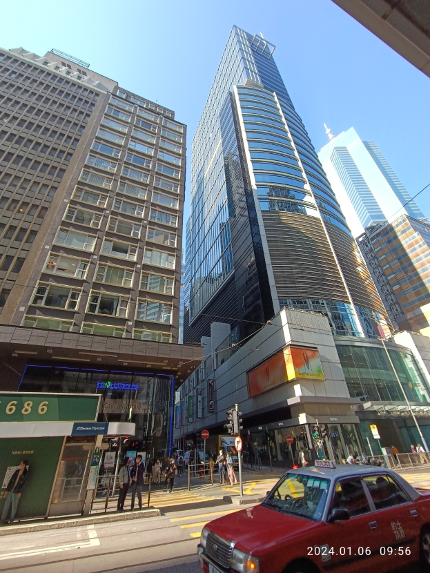 Ein rotes Auto fährt auf einer von hohen Wolkenkratzern gesäumten Stadtstraße, mit Passanten, Verkehrszeichen und Schildern, unter einem klaren Himmel.