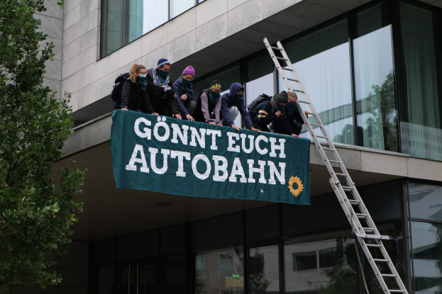 Gruppe von Menschen auf einem Gebäude mit einem Banner, einem Baum auf der linken Seite und Glasfenstern im Hintergrund, wahrscheinlich protestierend für die deutsche Regierung.
