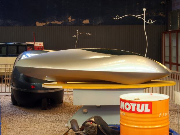 Kleines Hydrofoil-Boot auf einem Tisch neben einem Auto, mit zwei Trommeln auf dem Boden, einem Metallzaun und einer Glastür mit einer Tafel im Hintergrund.