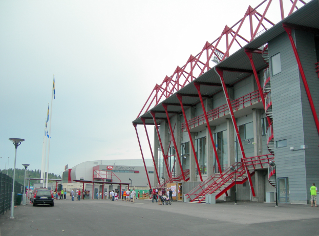 Ein großes Stadion mit Menschen, die herumlaufen, Fahrzeuge auf der Straße davor, ein Zaun auf der linken Seite und Bäume mit einem klaren blauen Himmel im Hintergrund.