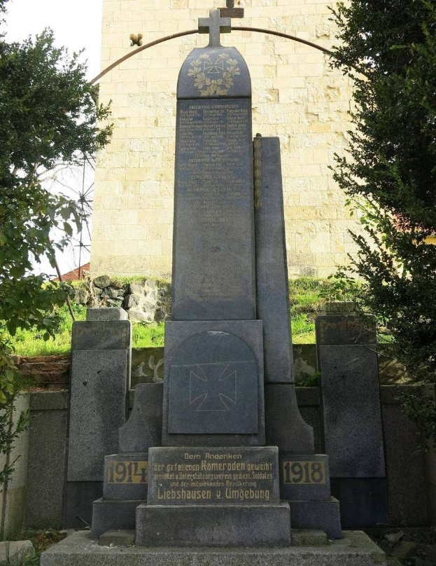 Ein steinernes Denkmal mit einem Kreuz, gewidmet den deutschen Soldaten, die im Zweiten Weltkrieg starben, steht vor einer Kirche mit einem Kreuz auf dem Dach, umgeben von Bäumen und einer Mauer.