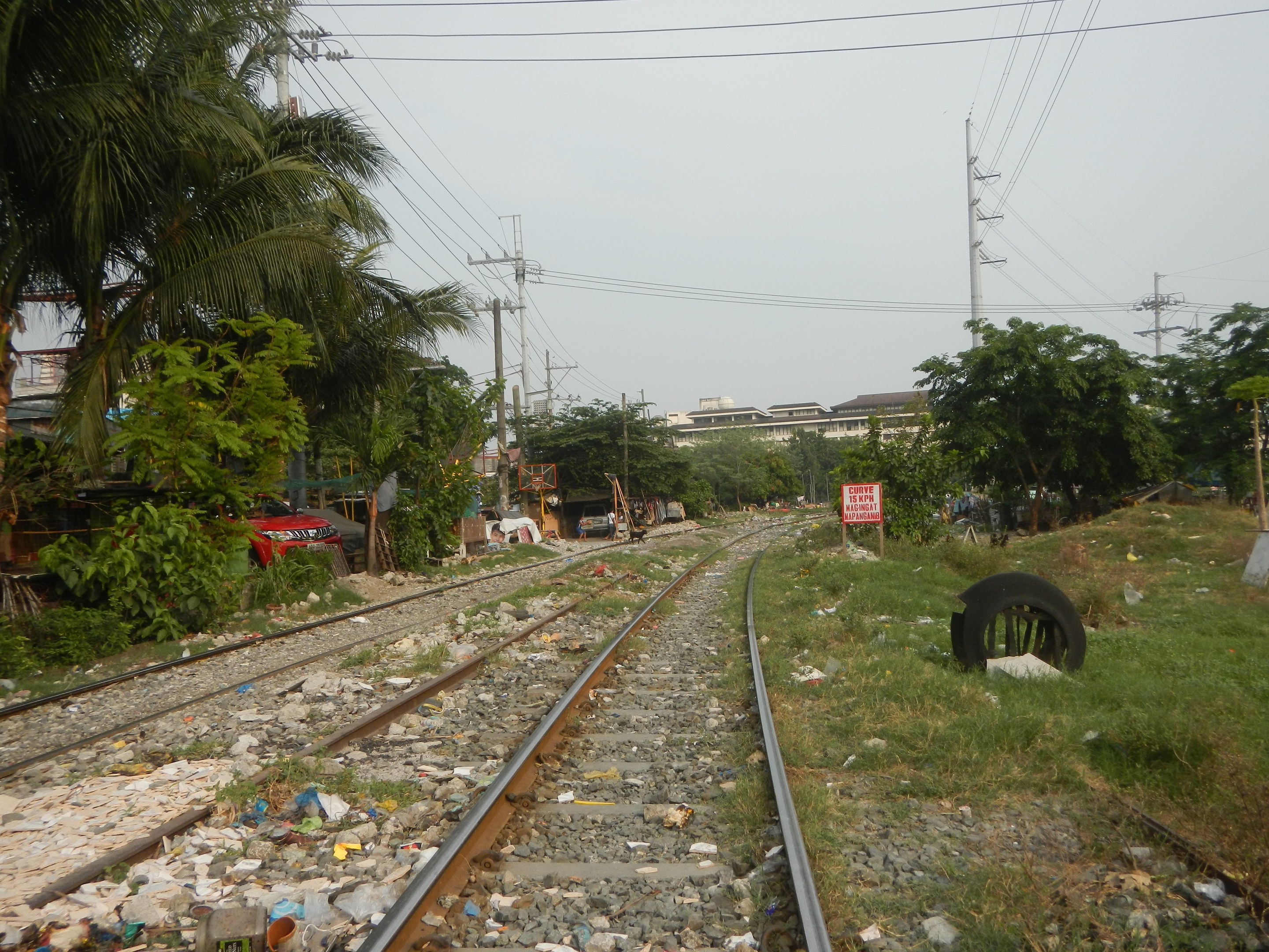 Eine Schienenstrecke mit einem Haufen Müll an der Seite, umgeben von Steinen, Gras, einem Reifen, einer Tafel mit Text, Bäumen, Strommästen mit Drähten, Fahrzeugen, Gebäuden und einem bewölkten Himmel.