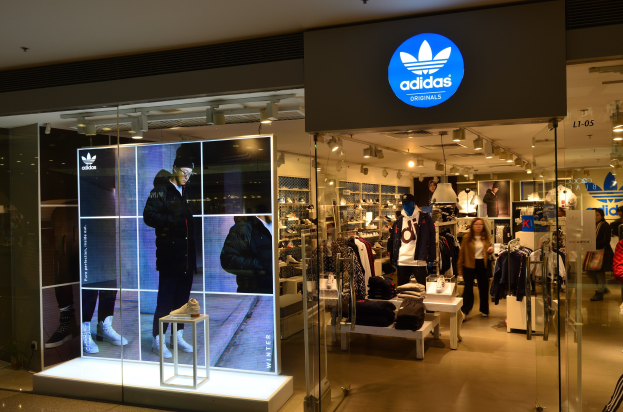 Außenansicht eines Adidas Originals-Ladens in einem Einkaufszentrum mit Schaufensterpuppen, Kleiderständern, Schuhen auf Ständern und beleuchteter Markenwerbung.