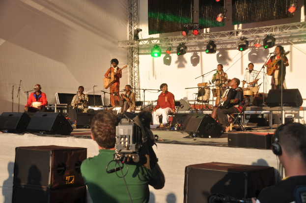 Ein musikalisches Konzert mit Musikern, die verschiedene Instrumente auf der Bühne spielen, darunter Gitarren und Trommeln, unter bunten roten und grünen Scheinwerfern, mit Lautsprechern vorne und zwei Personen unten, eine filmt und die andere lauscht.