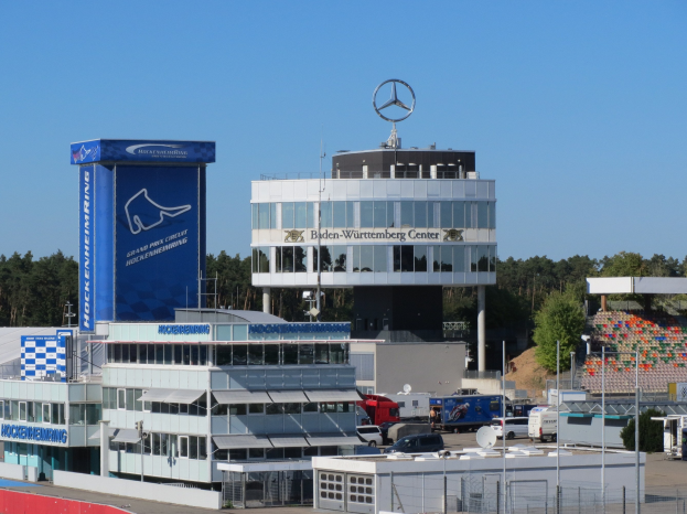 Übersicht des Mercedes-AMG Petronas F1 Team Headquarters in Berlin, Deutschland, mit Gebäuden, Fahrzeugen, einem Zaun, Beschriftungen, Bäumen und dem Team-Logo.