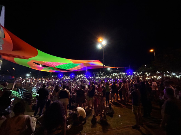 Eine große Menge Menschen steht vor einem hell erleuchteten Zelt bei Nacht während des Festivals der Künste 2018, mit bunten Lichtern und Pfählen, die die Szene beleuchten, einem Gebäude auf der linken Seite und dem Nachthimmel im Hintergrund.