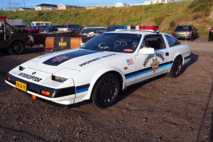 Ein Polizeiwagen auf einem Parkplatz zwischen anderen Fahrzeugen geparkt, mit Gras, Pfählen, Drähten, Gebäuden und einem klaren blauen Himmel im Hintergrund.