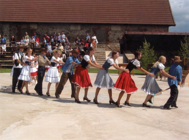 Eine Gruppe von Menschen in traditioneller bayrischer Tracht tanzt vor einem Gebäude mit Dach und Fenstern, einige tragen Hüte und halten Stöcke, während andere auf Bänken sitzen, mit Gras unter den Füßen und Bäumen im bewölkten Himmel im Hintergrund.