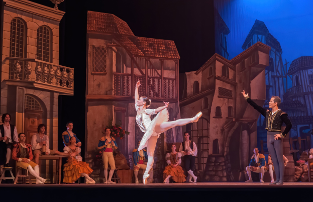 Zwei Tänzer performen auf einer Bühne im Royal Opera House in London, mit einem Publikum im Vordergrund, das sitzt und steht, und einem Bildschirm im Hintergrund, der Gebäude anzeigt.