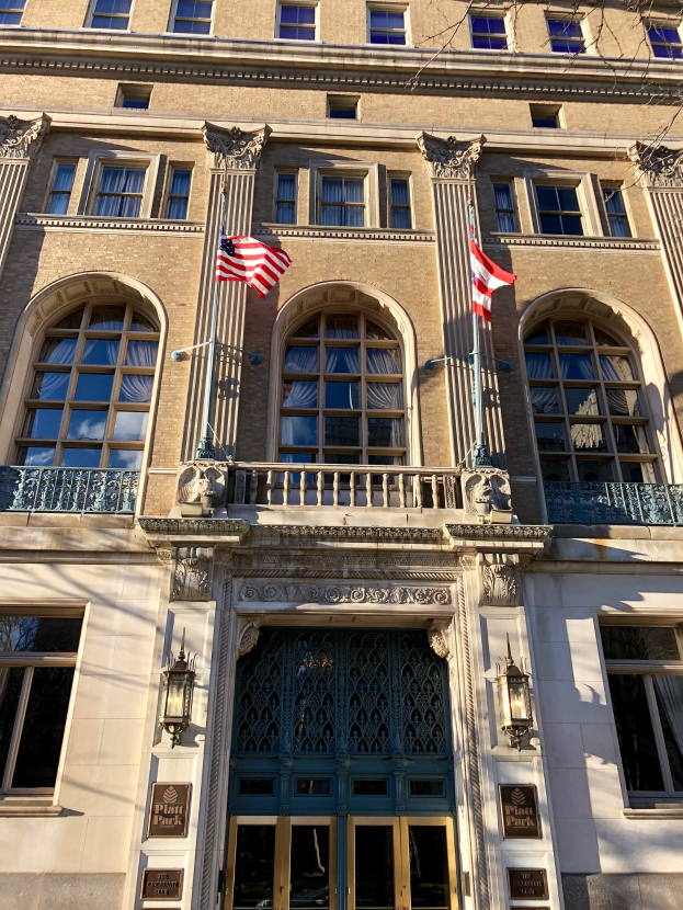 Außenansicht des Plaza Hotels in New York City, mit Glasfenstern, -türen, -säulen, -geländern, -leuchten, Texttafeln und Flaggen vor einem Himmelhintergrund.