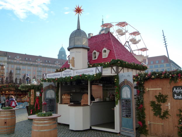 Ein kleines Gebäude mit einem roten Dach und einem Riesenrad davor, umgeben von Festdekorationen und Menschen, mit Gebäuden, Bäumen und einem bewölkten Himmel im Hintergrund.