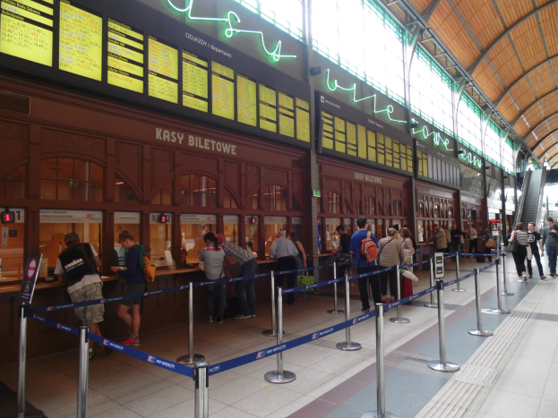 Menschen stehen in einer Warteschlange an einem Bahnhofsschalter mit Taschen, Stangen mit Bändern, Informationsschildern, Deckenleuchten und Treppen mit Geländern.