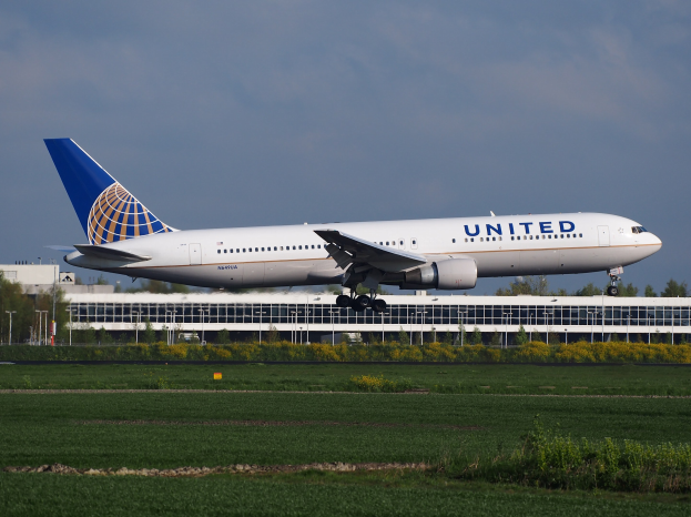 United Airlines Boeing 787-9 Dreamliner startet von einer Rollbahn mit grünem Gras, Bäumen, einem Gebäude und einem klaren blauen Himmel im Hintergrund.