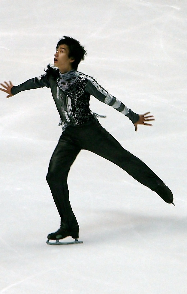 Männliche Eiskunstläufer in schwarzer Kleidung gleitet über eine Eisfläche und führt eine Choreographie mit synchronen Arm- und Beinbewegungen aus.