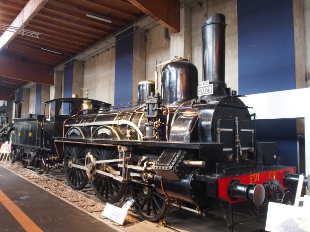 Eine gro├če, alte Dampflokomotive in einem Museums Setting mit umliegenden Eisenbahnschienen, Informationsschildern und anderen Ausstellungen.