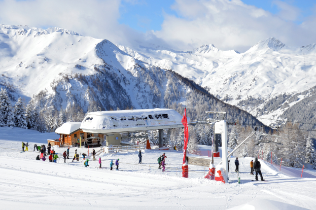 Gruppe von Skifahrern, die eine schneebedeckte Abfahrt im Österreichischen Alpengebiet hinunterfahren, mit Bäumen, Pfählen, Flaggen und einer Hütte im Hintergrund, vor einem bewölkten Himmel und einer montagneusen Kulisse.