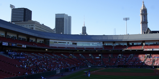 Ein überfülltes Baseball-Stadion mit Zuschauern, die ein Spiel beobachten, Spielern auf dem Feld und im Hintergrund Gebäude, Laternenmasten und einen klaren blauen Himmel.