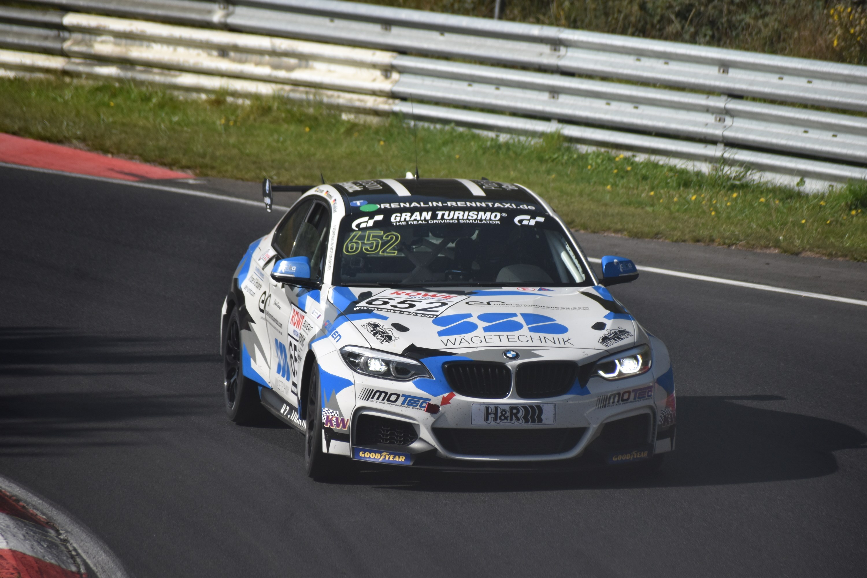 Ein BMW M4 DTM-Rennwagen mit Text und Zahlen auf der Karosserie, der auf einer von Gras, einem Zaun und Pflanzen umgebenen Rennstrecke fährt, wobei ein Rad sichtbar ist.