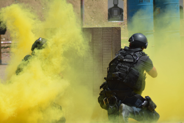 Zwei Polizisten in Einsatzausrüstung atmen gelben Rauch aus, tragen Helme und Schutzausrüstung, mit einer verbarrikadierten Wand und sichtbarem Boden im Hintergrund.