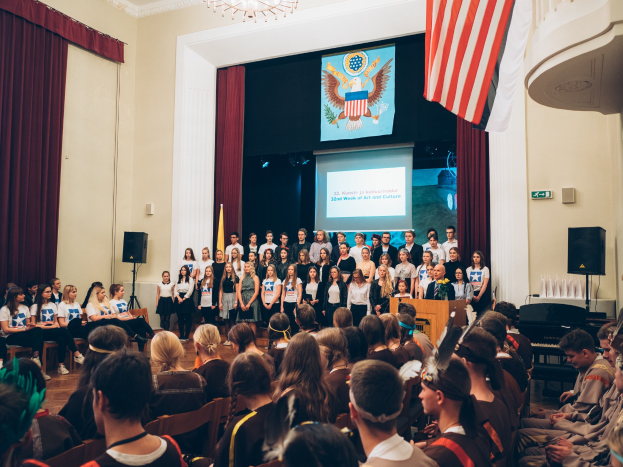 Eine Gruppe von Menschen steht vor einem sitzenden Publikum auf einem Podium mit Mikrofon, flankiert von Fahnen und Lautsprechern, während eines 2. Kunst- und Kulturwochen-Events.
