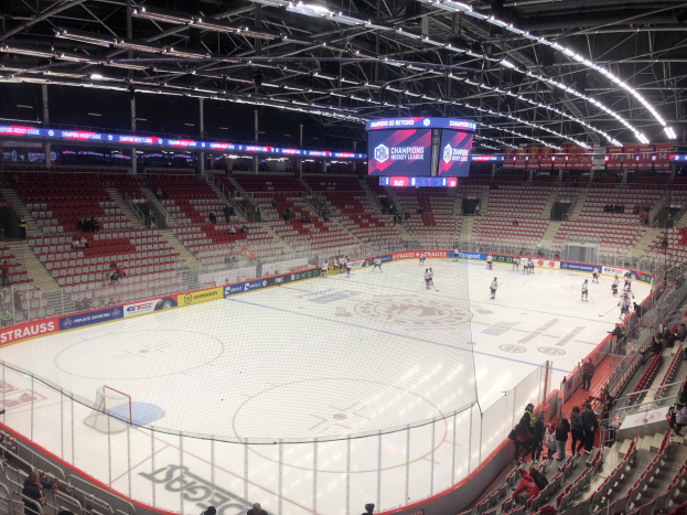 Eishockeyspiel in einer großen Arena mit Zuschauern, ein von einem Zaun umgebenes Spielfeld, ein Display mit der Aufschrift "St. Louis Blues gegen Detroit Red Wings", Metallrahmen, Deckenleuchten und ein Dach.