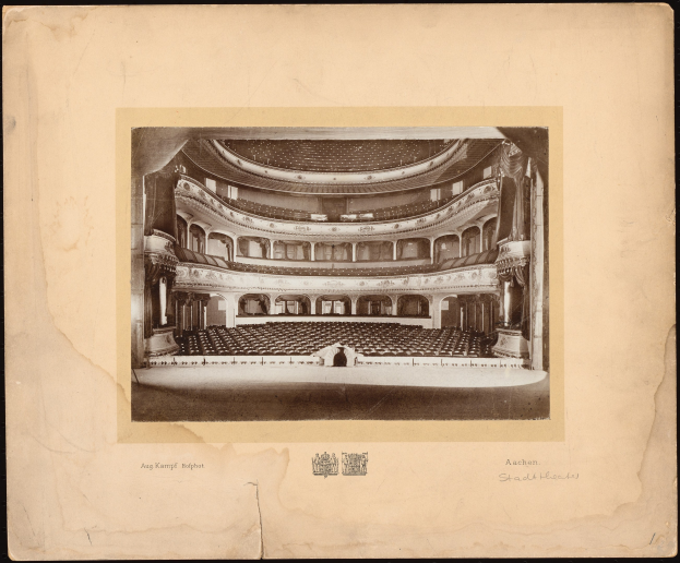 Schwarzes und weißes Foto des Auditoriums des Royal Albert Hall, das die Bühne, Reihen von Stühlen, Säulen, Geländer und Text mit einem Logo unten zeigt.