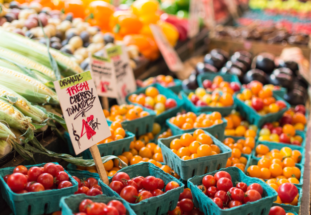 Ein Bauernmarkt mit Körben voller frischer Produkte, darunter Tomaten und Mais, sowie Schilder im Hintergrund.