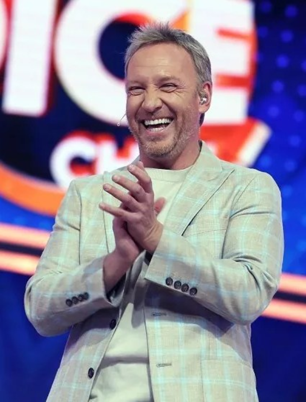 Ein Mann in einem Anzug klatscht auf der Bühne mit 'The Voice'-Logo im Hintergrund.