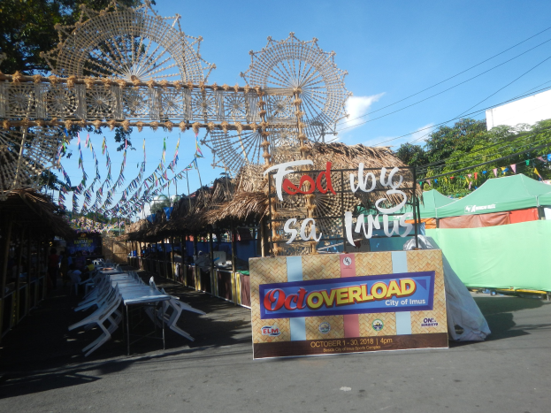Eine KarnevalsSzene mit einem großen Riesenrad in der Mitte, umgeben von Hütten, Sitzbereichen, einer Schautafel, Dekorationen, Bäumen, Überlandleitungen und einem bewölkten Himmel.