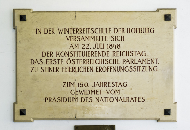 Plakette auf einem Gebäude mit der Aufschrift 'In der Winterreitschule der Hofburg' in schwarzer Schrift auf hellblauem Hintergrund mit schwarzem Rand.
