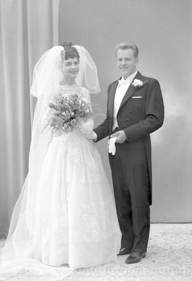 Schwarzes und weißes Foto eines Mannes und einer Frau, die nebeneinander auf ihrer Hochzeit stehen, die Frau in einem weißen Kleid mit einem Strauß Blumen, der Mann neben ihr mit einem Lächeln, mit einem Vorhang und einer Wand im Hintergrund.