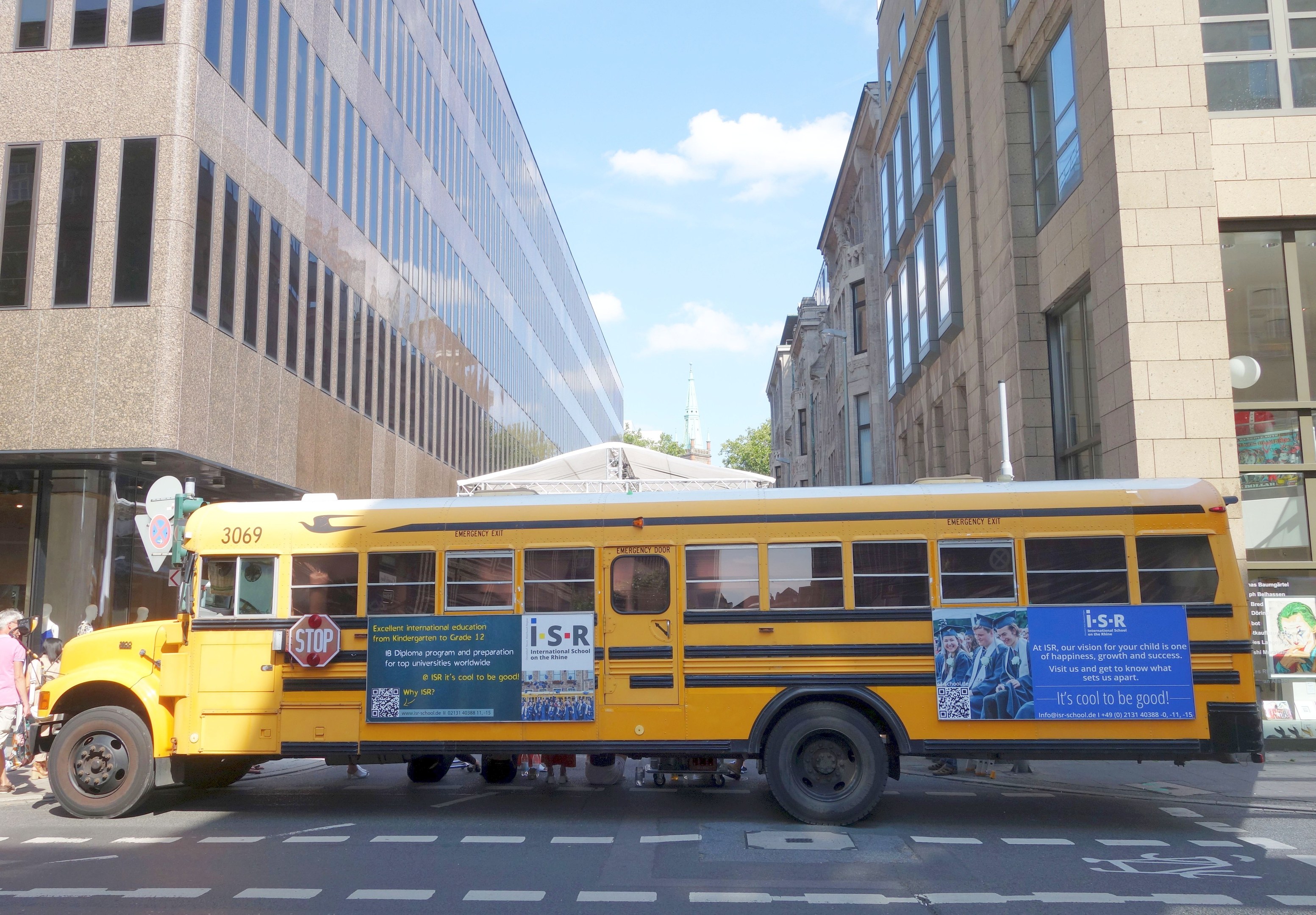 Ein gelber Schulbus fährt eine Straße entlang, die von hohen Gebäuden gesäumt ist, mit Passanten auf dem linken Gehweg, Bäumen im Hintergrund und einem klaren blauen Himmel.