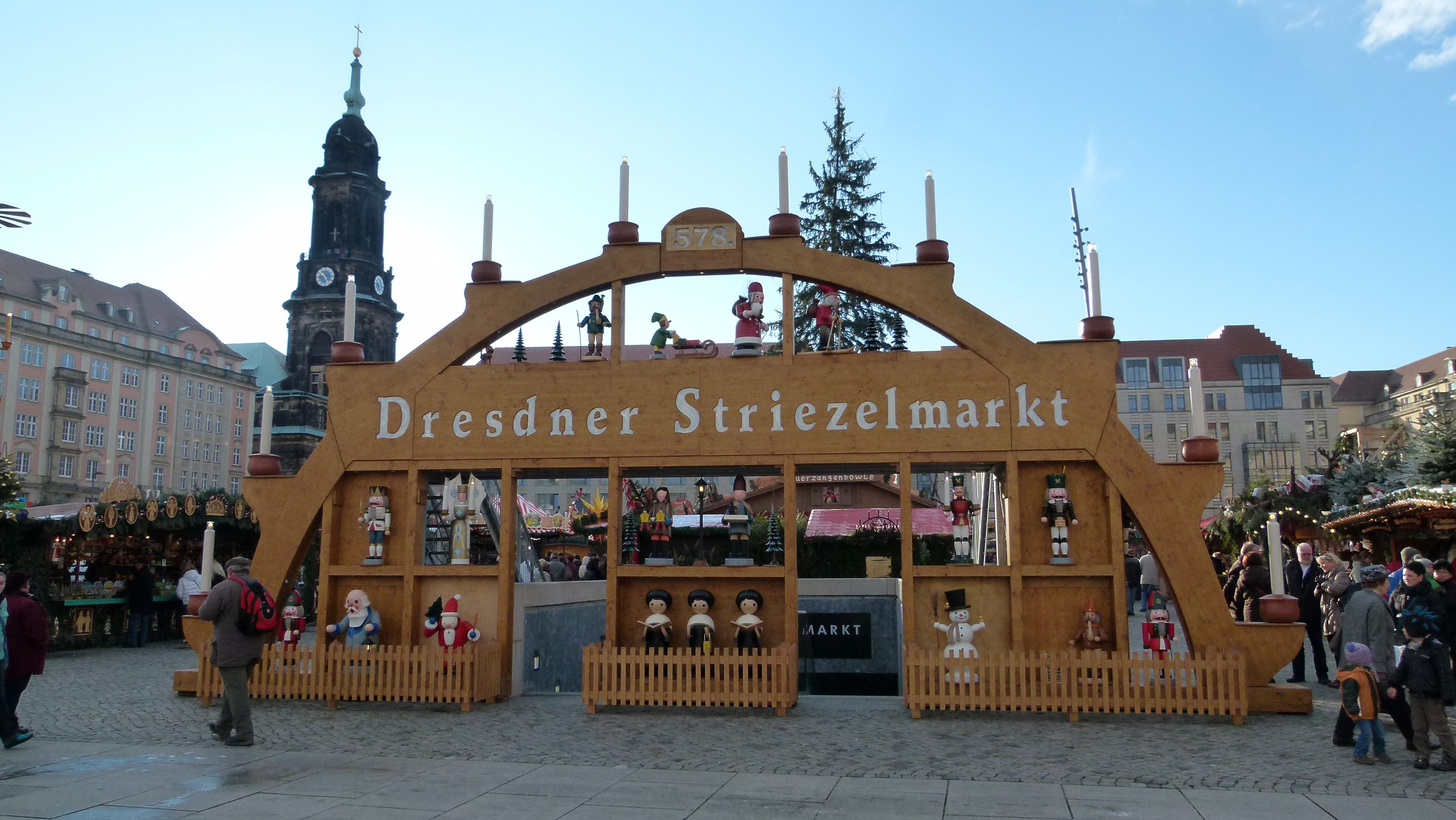 Ein belebter Weihnachtsmarkt in Dresden, Deutschland mit Menschen, die spazieren gehen, einem Torbogen, Bäumen, Gebäuden und einem bewölkten Himmel.