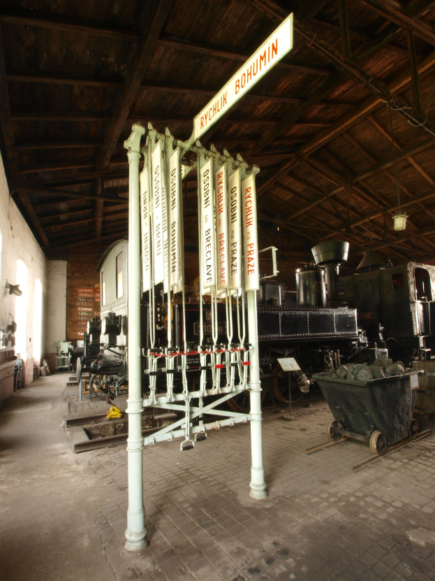 Ein Bahnsteig mit einem Zug auf den Schienen, einem Bodenwagen, einem an einem Pfahl befestigten Schild, einer Wand auf der linken Seite, einer Decke darüber und einer zentralen Signalbox.