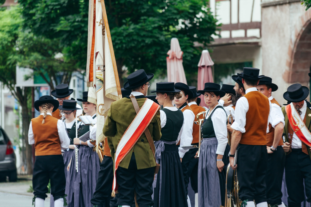 Eine Gruppe von Menschen in traditioneller bayrischer Tracht marschiert durch eine Straße, einige halten Musikinstrumente und Fahnen, im Hintergrund sind Bäume, Gebäude und ein Auto zu sehen.
